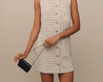 Peut inclure: Une robe sans manches à carreaux blancs et noirs avec un devant boutonné et une petite poche sur le côté gauche. La robe est associée à un sac à main noir à bandoulière chaîne et à des bottes noires mi-hautes.
