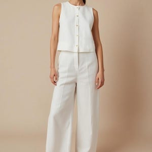 Pu&ograve; includere: Un top bianco senza maniche con bottoni dorati e pantaloni a gamba larga abbinati. L'outfit &egrave; realizzato in un tessuto colorato chiaro e ha un design minimalista. I pantaloni hanno una vita alta e una vestibilit&agrave; su misura, creando un look pulito ed elegante.