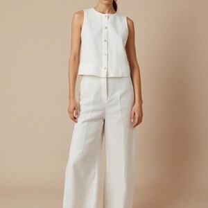 Pu&ograve; includere: Un top bianco senza maniche con bottoni dorati e pantaloni a gamba larga abbinati. L'outfit &egrave; realizzato in un tessuto strutturato e abbinato a ballerine nere. L'ensemble &egrave; in stile monocromatico e minimalista.