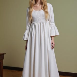 May include: A long, white dress with a square neckline and long sleeves with flared cuffs. The dress features a detailed bodice and a ruffled hemline. The model is wearing the dress in a room with a wooden table.