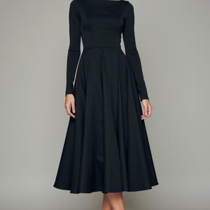 May include: A black long-sleeved midi dress with a boat neckline and a full skirt. The dress is fitted through the bodice and flows out into a wide, A-line skirt. The model is wearing the dress with nude heels.