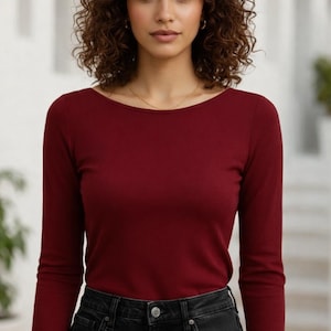 May include: A long-sleeved, burgundy-colored shirt with a scoop neckline. The shirt is paired with black denim jeans. The model is wearing a gold necklace and earrings. The background is a blurred outdoor setting.