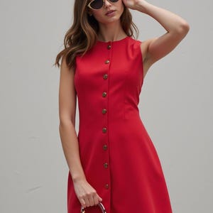 Può includere: Un abito rosso senza maniche con chiusura a bottoni sul davanti e un corpetto aderente. L'abito ha una gonna svasata e viene indossato con una piccola borsa a mano rosa.