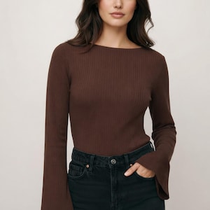 May include: A brown, ribbed top with a boat neck and flared sleeves, paired with black denim jeans. The top is long-sleeved. The model is wearing the top and jeans.