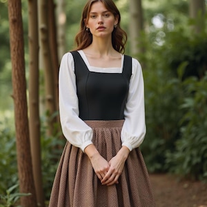 Puede incluir: Una mujer con un corpiño negro, una blusa blanca y una falda marrón con estampado de cuadros. Está de pie en un entorno de bosque.