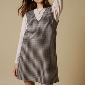 Könnte beinhalten: Ein braun-beiges Gingham-Kleid mit V-Ausschnitt, getragen über einem cremefarbenen Langarmshirt. Das Kleid hat eine einfache A-Linien-Silhouette. Das Model trägt eine goldene Halskette.