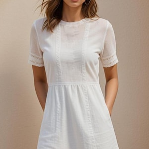 Könnte beinhalten: Ein weißes Baumwollkleid mit kurzen Ärmeln und einem Spitzenbesatz. Das Kleid hat einen runden Ausschnitt und einen leicht ausgestellten Rock.