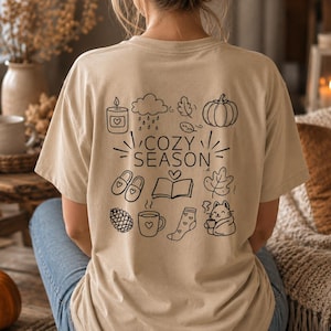 Könnte beinhalten: Ein beiges T-Shirt mit einer schwarzen Strichgrafik auf der Rückseite. Das Design enthält den Text "COZY SEASON", umgeben von Symbolen einer Kerze, einer Regenwolke, eines Kürbisses, Hausschuhen, einem offenen Buch, Blättern, einem Tannenzapfen, einer Tasse, einer Socke und einer Katze.