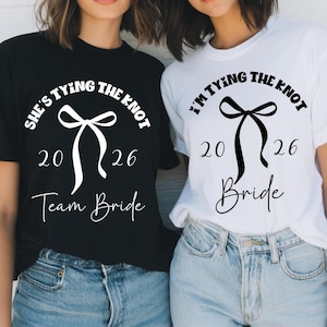 Puede incluir: Dos camisetas, una negra y otra blanca, con gráficos temáticos de boda. La camiseta negra dice "She's Tying the Knot", "2026" y "Team Bride". La camiseta blanca dice "I'm Tying the Knot", "2026" y "Bride".
