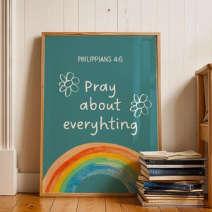 Può includere: Una stampa turchese incorniciata con il testo "Filippesi 4:6 Pregate per ogni cosa" e un'illustrazione arcobaleno. Due disegni di fiori bianchi sono su entrambi i lati del testo. La stampa è esposta su un pavimento in legno accanto a una pila di libri.