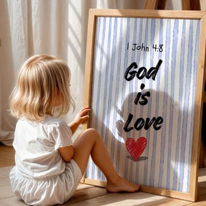 Può includere: Un'opera d'arte incorniciata con la scritta "God is Love" e un cuore rosso, su uno sfondo a righe blu e bianche. Un bambino è seduto sul pavimento, che guarda l'opera d'arte. La cornice è in legno chiaro.