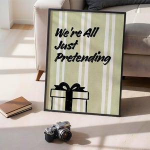 May include: A framed print with the text "We're All Just Pretending" above a gift box illustration. The background has vertical stripes in light green and white. A closed book and a camera are in the foreground.