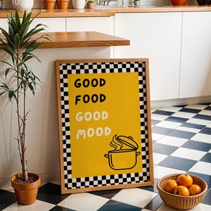 May include: A framed poster with the words "GOOD FOOD GOOD MOOD" in black and white checkered border. A yellow background features a pot illustration. A small potted plant and a basket of oranges are also in the image.
