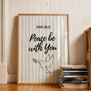 Può includere: Una stampa incorniciata con il testo "Peace be with You" e "John 20:21" su sfondo a righe. È presente anche una colomba che porta un ramoscello d'ulivo. La cornice è in legno e sotto c'è una pila di libri.