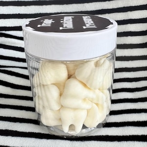 Peut inclure: Un pot en plastique transparent rempli de bonbons blancs en forme de dents. Le pot a un couvercle blanc avec les mots "Tantalizing Treasures" imprimés en noir. Le pot est posé sur un fond à rayures noires et blanches.