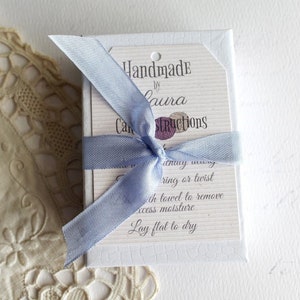 May include: A white box with a blue ribbon tied around it. The box has a tag attached to it that says "Handmade by Laura" and "Care Instructions". The instructions say to "lay flat to dry" after washing.