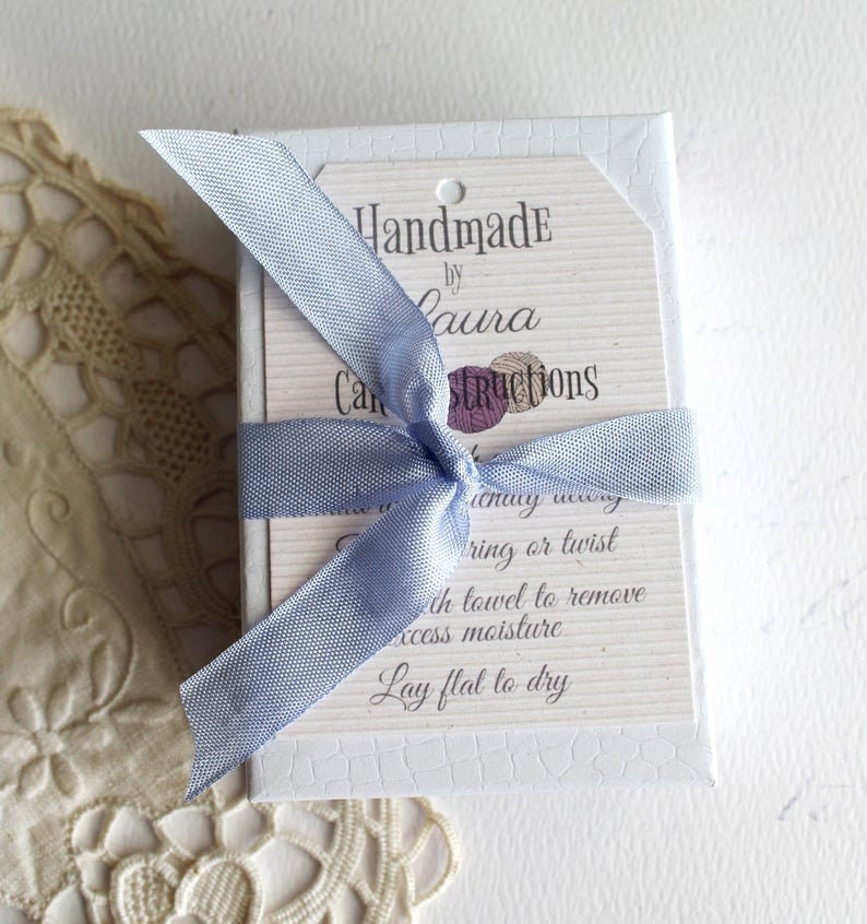May include: A white box with a blue ribbon tied around it. The box has a tag attached to it that says "Handmade by Laura" and "Care Instructions". The instructions say to "lay flat to dry" after washing.