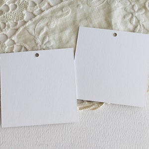 May include: Two blank white gift tags with a hole punched in the top corner. The tags are on a white surface with a white lace doily underneath.