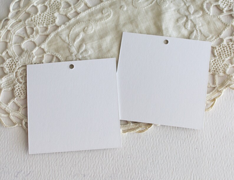 May include: Two blank white gift tags with a hole punched in the top corner. The tags are on a white surface with a white lace doily underneath.