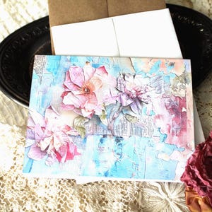 May include: A greeting card with a floral design. The card features a collage of pink and purple flowers against a blue and white background. The card is accompanied by a brown envelope and a white envelope.