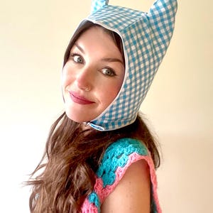 May include: A blue and white gingham bonnet with pointed ears. The bonnet is worn by a person with long brown hair and a pink and blue crocheted top.