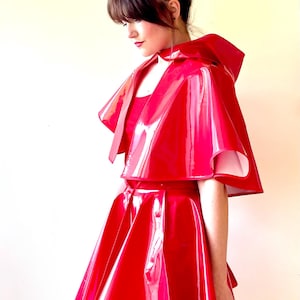 Könnte beinhalten: Ein roter Vinylrock und ein passendes Cape mit Kapuze. Der Rock ist A-förmig und das Cape hat einen breiten Kragen.