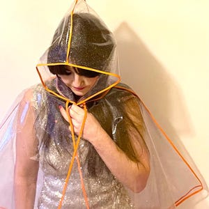 May include: A clear plastic raincoat with an orange trim. The raincoat has a hood and is worn over a silver sequined dress.