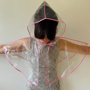 May include: A clear plastic hooded cape with a pink trim. The cape is worn over a sequined dress.