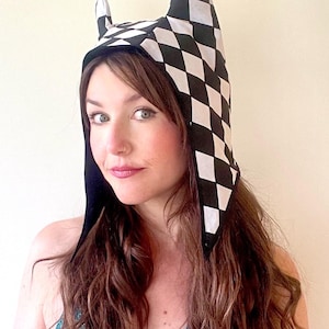 May include: A black and white checkered hooded hat with pointed ears. The hat is made of fabric and has a black lining. The hood is pulled up over the head, and the ears are pointing up.