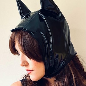 May include: A black, glossy, hooded head covering with pointed ears. The head covering is made of a shiny material and is worn by a person with long brown hair. The hood covers the head and neck.