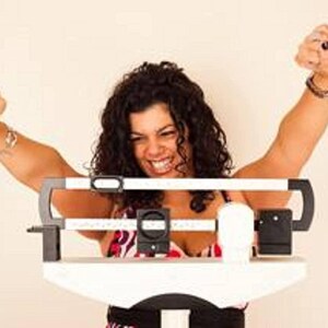 May include: A woman stands on a white digital scale with her arms raised in the air. She is smiling and appears happy.