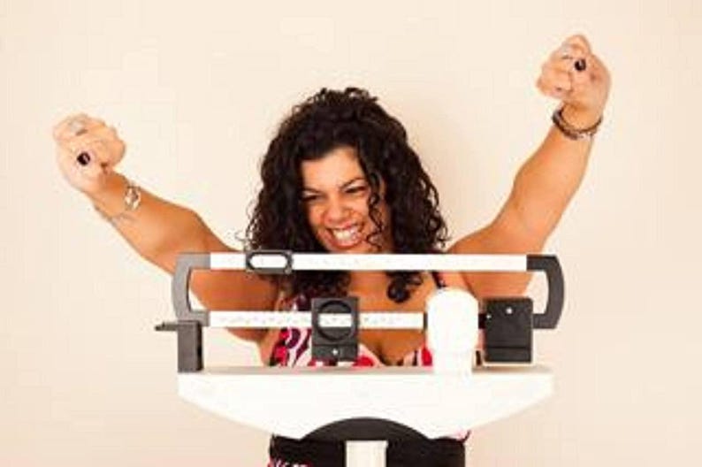 May include: A woman stands on a white digital scale with her arms raised in the air. She is smiling and appears happy.