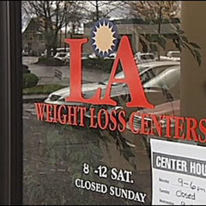May include: A storefront window with a red and white sign that reads "LA Weight Loss Centers". The sign has a yellow sun with rays in the center. The window also has a sign that says "Center Hours" and lists the hours of operation.