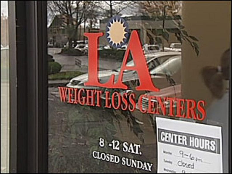 May include: A storefront window with a red and white sign that reads "LA Weight Loss Centers". The sign has a yellow sun with rays in the center. The window also has a sign that says "Center Hours" and lists the hours of operation.