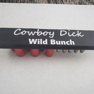 May include: Black wooden ammo holder with the text "Cowboy Dick Wild Bunch" in white lettering. The holder has eight red shotgun shells.