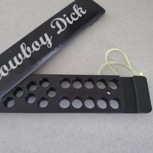 May include: A black metal bullet holder with a hinged lid. The lid has the words "Cowboy Dick" printed in white lettering.
