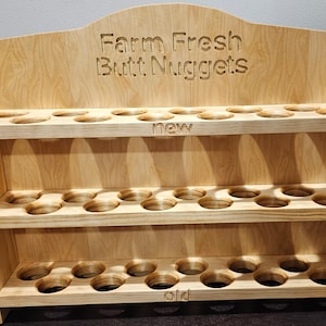 Puede incluir: Un expositor de madera con tres estantes, cada uno con múltiples recortes circulares. El estante superior tiene el texto "Farm Fresh Butt Nuggets" tallado en la madera. El estante del medio tiene la palabra "new" tallada en la madera. El estante inferior tiene la palabra "old" tallada en la madera.