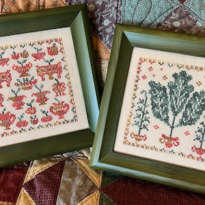 May include: Two framed cross-stitch pieces with an autumn theme. The first piece features a repeating pattern of apples, teapots, and small animals. The second piece features a large green plant with smaller plants on either side. The frames are a dark green wood.