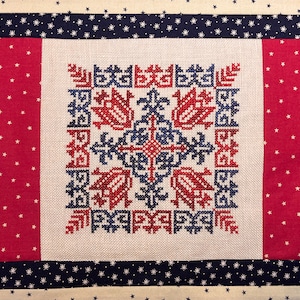 May include: A red, white and blue cross-stitch pattern on a beige fabric background. The pattern features a geometric design with stylised flowers and leaves.