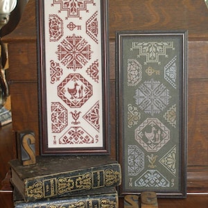 May include: Two framed cross-stitch embroidery pieces with a brown and a green background. The embroidery designs feature geometric patterns and a bird motif. The frames are on a wooden surface with books and letterpress blocks.