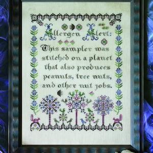 May include: A framed cross-stitch sampler with a white background and colorful embroidery. The text reads "Allergen Alert: This sampler was stitched on a planet that also produces peanuts, tree nuts, and other nut jobs."