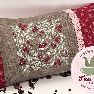 May include: A brown linen pillow with a red and white cross-stitch design of a teacup and berries. The pillow has a red and white gingham border and a pink lace trim. The pillow is sitting on a white surface with loose tea leaves scattered around it. A pink circle with the text "Treenway Silks Tea Time Palette @xstitchtherainbow" is in the lower right corner of the image.