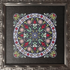 May include: A framed cross-stitch featuring a symmetrical floral design. The artwork showcases a central starburst pattern in teal, red, and green, surrounded by a circular border with small pink and blue floral motifs. The frame is a metallic silver color.