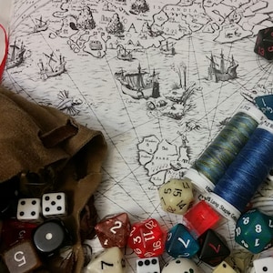 May include: A collection of dice, a leather pouch, and spools of thread on a vintage map. The dice are various colors, including red, blue, green, and black. The map is a black and white illustration of a sea with ships and islands.