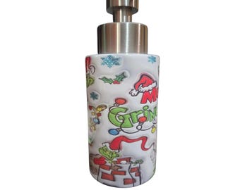 May include: A cylindrical white soap dispenser with a brushed metal pump. The surface features a colorful holiday-themed print including a green character, red Santa hats, holly, snowflakes, and the text Merry Grinchmas in green and red.