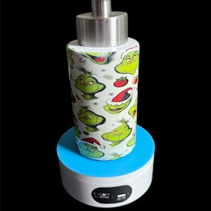May include: A white and silver liquid soap dispenser with a repeating pattern of the Grinch's face in green, with red and white Santa hats and ornaments. The dispenser has a silver pump and is displayed on a blue and white rotating stand.