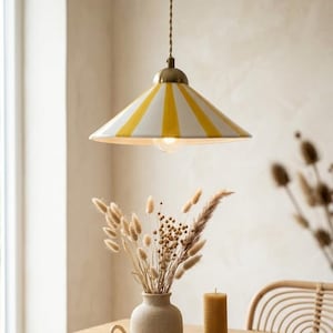 Può includere: Una lampada a sospensione con un paralume a righe bianche e gialle. La lampada ha una parte superiore color ottone e un cavo marrone intrecciato. La luce è sospesa sopra un tavolo con un vaso di fiori secchi e una candela.