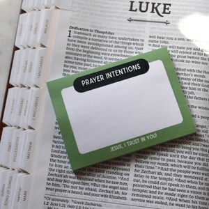 May include: Open Bible with a green and white card that reads "Prayer Intentions" and "Jesus, I trust in You!" The Bible has white tabs and is open to the book of Luke.