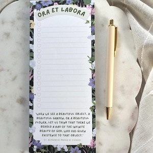 May include: A floral-bordered notepad with the text "ORA ET LABORA" and lined pages for lists. A quote by St. Alphonsus Maria de Liguori is printed at the bottom. A cream-colored pen with gold accents rests beside the notepad.