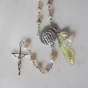 May include: A rosary necklace featuring off-white beads, a silver-toned crucifix, and a central circular medal depicting a face. The piece includes a dangling charm set with a translucent pale green leaf-shaped pendant and a small white bead.